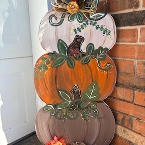 May include: A wooden, three-tiered pumpkin decoration. The top pumpkin is white, the middle is orange, and the bottom is brown. Each pumpkin is decorated with green leaves and vines. The top is adorned with a bow and small pumpkins. A burlap and leaf base completes the fall-themed decor.