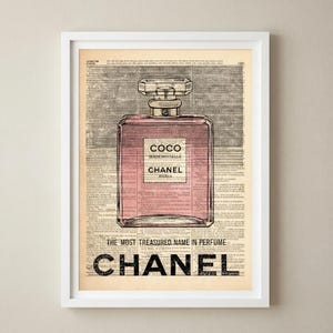 May include: Framed print featuring a pink perfume bottle with the text "COCO MADEMOISELLE CHANEL PARIS" on a vintage newspaper background. The bottom of the print reads "THE MOST TREASURED NAME IN PERFUME" and "CHANEL".