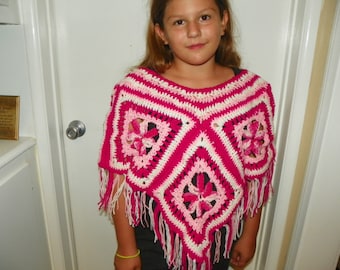 Op de afbeelding: Een roze en witte gehaakte poncho met een bloemenpatroon en franjes. De poncho wordt gedragen door een jong persoon met lang bruin haar.