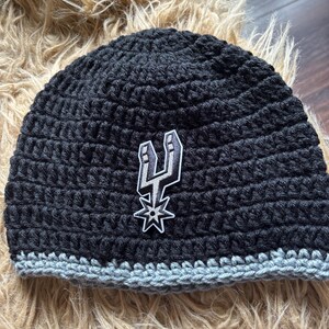 May include: A black crocheted beanie featuring a gray crocheted brim and a centered embroidered patch of the San Antonio Spurs basketball team logo, which depicts a stylized silver and black spur with a star shape at the bottom.