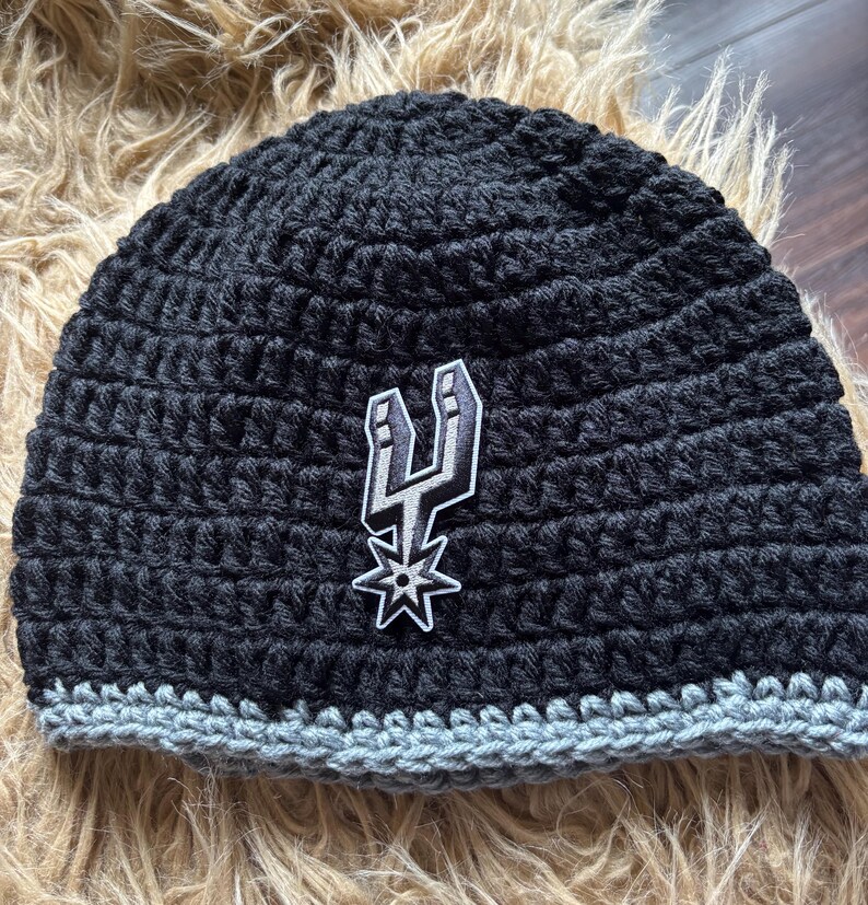 May include: A black crocheted beanie featuring a gray crocheted brim and a centered embroidered patch of the San Antonio Spurs basketball team logo, which depicts a stylized silver and black spur with a star shape at the bottom.
