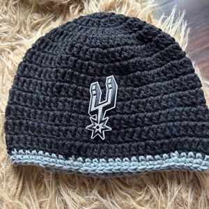 May include: A black crocheted beanie featuring a gray brim and a centered San Antonio Spurs logo patch. The hat is constructed with a thick yarn texture and displays the team's iconic spur emblem in black, white, and gray on the front panel.