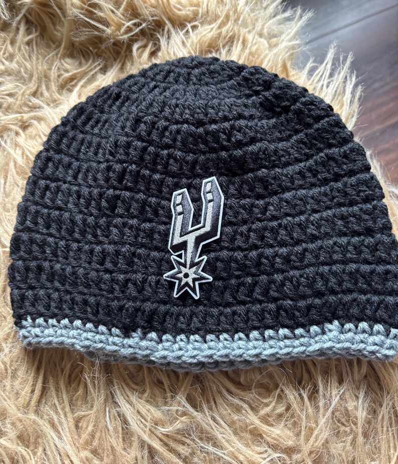 May include: A black crocheted beanie featuring a gray brim and a centered San Antonio Spurs logo patch. The hat is constructed with a thick yarn texture and displays the team's iconic spur emblem in black, white, and gray on the front panel.