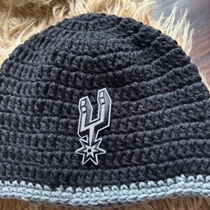 May include: A black crocheted beanie featuring a gray bottom border and a centered embroidered patch of the San Antonio Spurs basketball team logo in black, white, and gray. The hat is constructed with a thick, textured yarn stitch pattern.