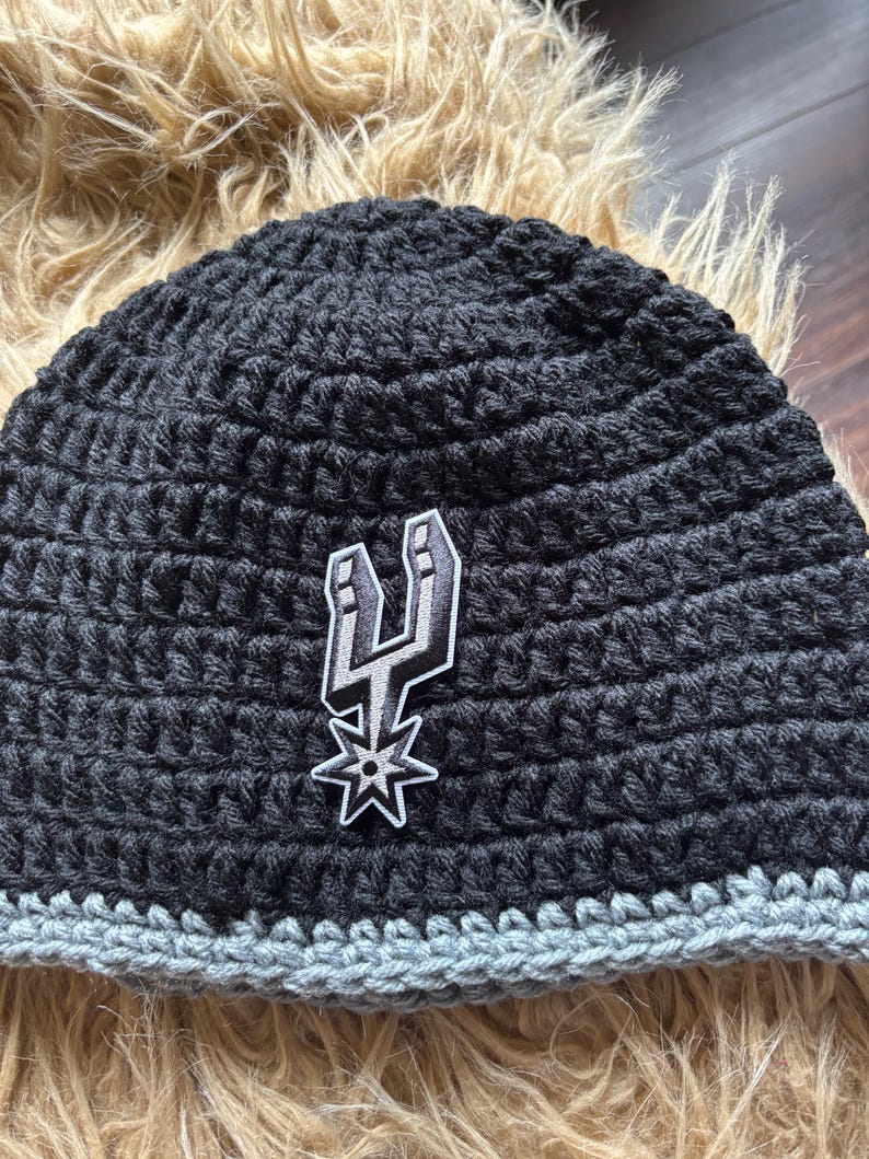 May include: A black crocheted beanie featuring a gray bottom border and a centered embroidered patch of the San Antonio Spurs basketball team logo in black, white, and gray. The hat is constructed with a thick, textured yarn stitch pattern.