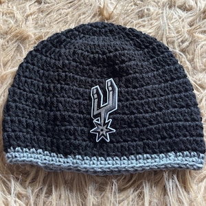 May include: A black crocheted beanie featuring a gray bottom border and a centered patch displaying the San Antonio Spurs logo in black, white, and gray. The hat is constructed with a textured yarn stitch pattern and a solid, rounded crown shape.