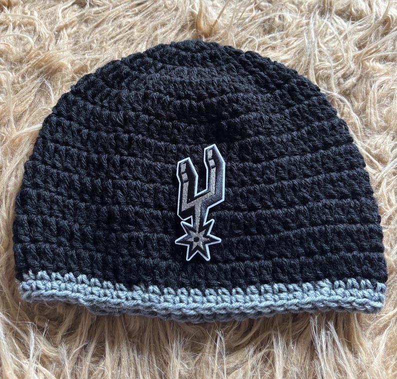 May include: A black crocheted beanie featuring a gray bottom border and a centered patch displaying the San Antonio Spurs logo in black, white, and gray. The hat is constructed with a textured yarn stitch pattern and a solid, rounded crown shape.