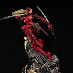 May include: A detailed figurine of a female superhero in a red and black costume, holding two katanas. The figure has blonde hair and is posed mid-leap on a rocky base. The statue includes a gun and is set against a dark backdrop.