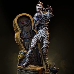 May include: A detailed figurine of a mummy, wrapped in white bandages, with a distressed expression. It stands before a golden sarcophagus and a statue of Anubis. A black dog statue is at the base. The figure is on a stone base.