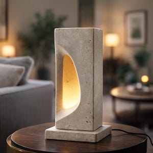 Pode incluir: Candeeiro de mesa retangular de pedra bege com um recorte curvo na frente. O candeeiro assenta numa base quadrada e emite um brilho quente e convidativo. O candeeiro está numa mesa castanha escura.