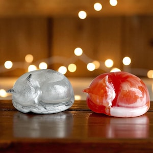 May include: Two glass fox ornaments, one silver and one red, are curled up asleep. The silver fox has a glittery finish, while the red fox has a marbled white pattern. They sit on a wooden surface with blurred lights in the background.