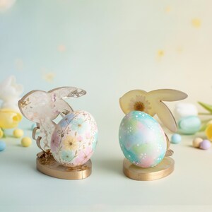 May include: Two Easter egg holders with bunny silhouettes. Each holder has a gold base and holds a decorative egg. One egg is decorated with pastel flowers, the other with a watercolor design. The background is a soft, blurred pastel.