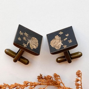 Puede incluir: Un par de gemelos cuadrados con fondo oscuro y diseños florales dorados. Los gemelos tienen forma rectangular con una barra dorada y un patrón floral decorativo. La estética general es vintage y elegante.