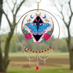 May include: A circular suncatcher featuring a blue moth design with pink roses, a black diamond, and gold accents. The suncatcher is suspended by a gold chain and adorned with red and green beads and a pink crystal pendant.