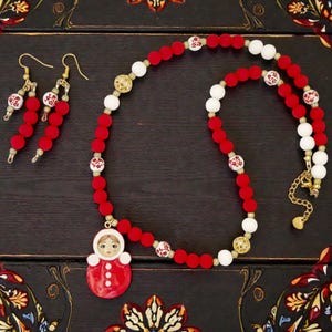 May include: A red beaded necklace and matching earrings set. The necklace features red and white beads, gold accents, and a pendant of a doll. The earrings have red beads. The jewelry is displayed on a dark wooden surface.