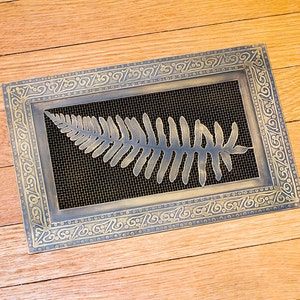 May include: A gold-colored metal vent cover with an ornate design featuring a fern leaf. The vent cover has a black mesh screen behind the fern.