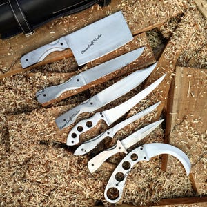 Puede incluir: Colección de cuchillos de acero inoxidable con mangos de madera y texto "Armor Craft Master" grabado en las hojas. Los cuchillos están dispuestos sobre una superficie de madera cubierta de virutas, con un estuche de cuero negro.