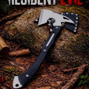 Pode incluir: Um machado preto e prateado com o texto "RESIDENT EVIL" em branco e vermelho. O machado tem um cabo preto e uma lâmina prateada. O machado está sobre uma rocha e rodeado por madeira e folhas.