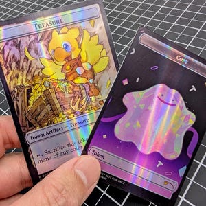 May include: Two rectangular trading cards with holographic finishes. The left card features a yellow bird character above a wooden treasure chest with the text Treasure and Token Artifact. The right card shows a pink blob character with the text Copy.
