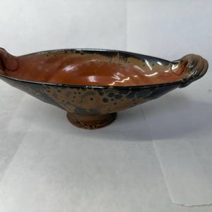 May include: A ceramic bowl with a pedestal base and two ornate handles. The interior is a warm orange, while the exterior features a speckled pattern of black and brown. The bowl's rim is dark, creating a contrast with the lighter interior.