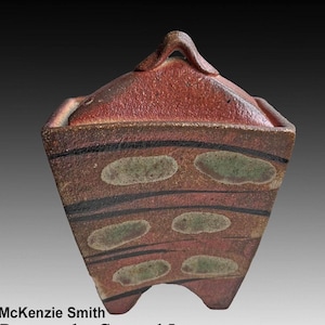 Puede incluir: Caja con tapa de cerámica artesanal con estética rústica. La caja presenta un exterior marrón rojizo con rayas negras horizontales y detalles ovalados verdes. La tapa tiene un asa y la caja descansa sobre pequeños pies. Una pieza de cerámica única.