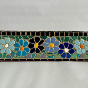 Könnte beinhalten: Ein rechteckiges Mosaikstück mit fünf stilisierten Blumen in Blautönen mit weißen und orangefarbenen Zentren, vor einem grünen und schwarzen Hintergrund. Das Mosaik ist mit schwarzen Fliesen umrandet.