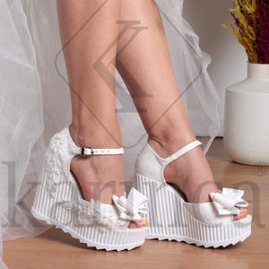 May include: White platform wedge sandals with a textured design and ankle straps. The sandals feature a decorative bow on the toe and are made of a textured fabric. The wedge heel has a ridged pattern.