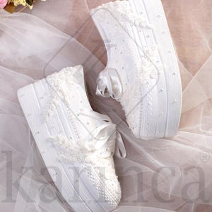 Pode incluir: Dois sapatos de noiva brancos, decorados com renda, fitas e strass. Estão sobre um tecido branco transparente, com um buquê de flores rosas e brancas ao fundo. Os sapatos têm uma superfície texturizada.