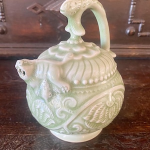 May include: A pale green ceramic teapot with a detailed design. The teapot features a curved handle, a spout with a sculpted animal head, and ornate relief patterns. The lid is topped with a decorative finial.