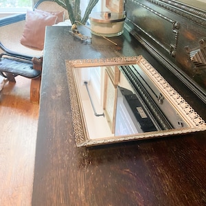 May include: A rectangular vanity mirror featuring a decorative, ornate gold-toned metal filigree border. The mirror has a flat, reflective glass surface with intricate metalwork detailing along all four edges, resting on a dark wooden surface.