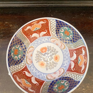 May include: A round ceramic plate featuring a central white medallion with orange floral motifs surrounded by alternating panels of deep red with white floral patterns and dark blue geometric grids containing green and orange circular designs.