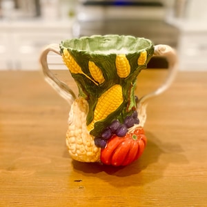 May include: A ceramic jug with a harvest theme. The jug features a green rim and is decorated with corn, grapes, and a red pumpkin. The handle is a light beige colour. The jug is sitting on a wooden surface.
