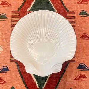 Könnte beinhalten: Eine weiße, muschelförmige Dekoschale. Die Schale hat eine gerippte Textur, die von der Mitte ausgeht. Die Schale liegt auf einem gemusterten Teppich mit roten, grünen und pfirsichfarbenen Tönen.
