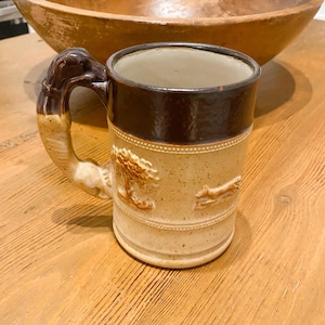 May include: A tan ceramic mug with a dark brown rim and handle. The handle is shaped like a stylized animal head. The mug features embossed images of a tree and a running animal. The mug is sitting on a wooden surface.