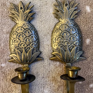 May include: Two brass pineapple-shaped wall sconces. Each sconce features a detailed pineapple design with a candle holder. The sconces are made of brass and have a vintage look. The pineapple design is a popular tropical decor element.