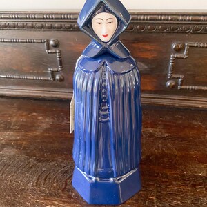 Könnte beinhalten: Blaue Keramikfigur einer Frau in einem Kapuzenmantel. Die Figur hat ein weißes Gesicht mit roten Lippen und steht auf einem blauen sechseckigen Sockel. Der Umhang hat eine geknöpfte Vorderseite.