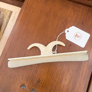 Peut inclure: Une brosse à vêtements blanc cassé dotée d'une poignée incurvée avec un motif en relief et une bande de nettoyage en tissu doux de couleur claire. Une étiquette en papier blanc est attachée par une ficelle, portant le texte THE ENGLISH COBBLER.