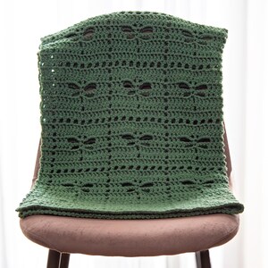 May include: A handmade, forest green crochet blanket draped over a chair. The blanket features a repeating pattern of openwork butterfly shapes and horizontal rows. The blanket is made of soft yarn and is a cozy home decor item.