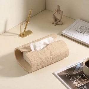 May include: A beige, textured tissue box with a curved design, holding white tissues. A gold pen holder and a small statue are also on the table. A magazine and a cup of coffee are also visible.