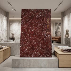 Pode incluir: Uma grande laje retangular de mármore vermelho com veios brancos, exibida em um showroom. A pedra polida está em pé sobre uma base de concreto, mostrando seus padrões e texturas naturais. O showroom apresenta outras amostras de mármore e esculturas.