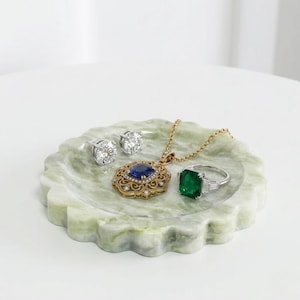 May include: Jewellery collection on a green and white marble tray. Includes diamond stud earrings, a gold pendant with a blue gemstone, a silver ring with a green gemstone, and a gold necklace.