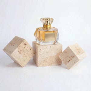 May include: A clear glass perfume bottle with a gold cap and a gold label, filled with a light amber liquid. The bottle is displayed on a beige stone cube, with two additional stone cubes on either side.