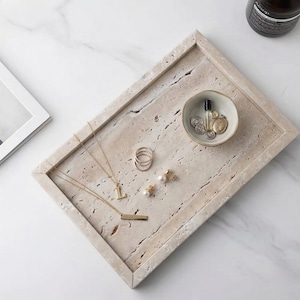 May include: A rectangular, beige stone tray holds various jewelry items. Gold necklaces, rings, and earrings are arranged alongside a small ceramic bowl containing coins and a perfume bottle. A dark bottle is visible in the background.