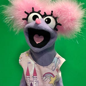 May include: A purple hand puppet with large white eyes, black eyelashes, and a small pink nose. The puppet has two fluffy pink pom-poms on its head and wears a white dress featuring a colorful pattern of castles, rainbows, stars, and unicorns.