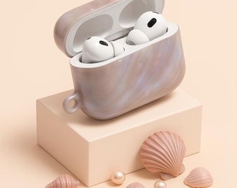 May include: A marbled, pearlescent case for wireless earbuds, open to reveal two white earbuds. The case sits on a light peach-coloured block, with decorative seashells and pearls scattered around it.
