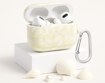 Może przedstawiać: Jasnożółte etui na AirPods Pro z pikowanym wzorem, otwarte, aby odsłonić białe słuchawki. Dołączony jest srebrny karabińczyk. Etui stoi na białym bloku, z białymi muszlami i perłami ułożonymi poniżej.