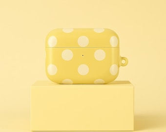 Puede incluir: Una funda amarilla para AirPods con un estampado de lunares blancos. La funda es rectangular con bordes redondeados y un pequeño lazo lateral. Se apoya sobre un bloque rectangular amarillo a juego.