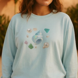 May include: A light blue sweatshirt featuring a seashell and pearl graphic. The design includes a large, iridescent blue seashell, scattered pearls, and colorful sea glass shapes. The sweatshirt is a solid light blue color.