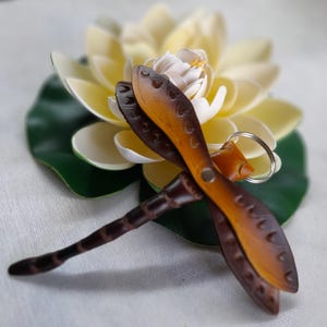 May include: A handcrafted leather dragonfly keychain with brown and tan wings and a dark brown body. The dragonfly is attached to a silver keyring. The dragonfly rests on a white and yellow flower with a green leaf.