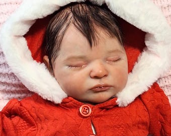 May include: A realistic baby doll with closed eyes and dark hair, wearing a red textured long-sleeve garment with two red buttons and a hood trimmed with white faux fur. The doll has a fair complexion and is positioned in a resting pose.